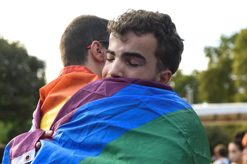 Two boys embrace, with their eyes closed, while wrapped in a rainbow flag. 