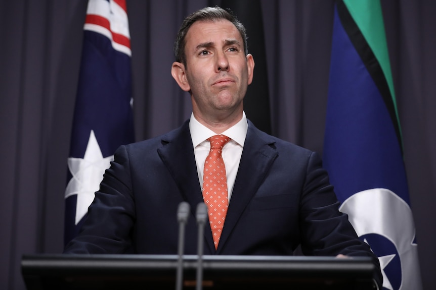 Jim Chalmers stands behind a podium and in front of the Australian and Torres Strait Islander flags. 