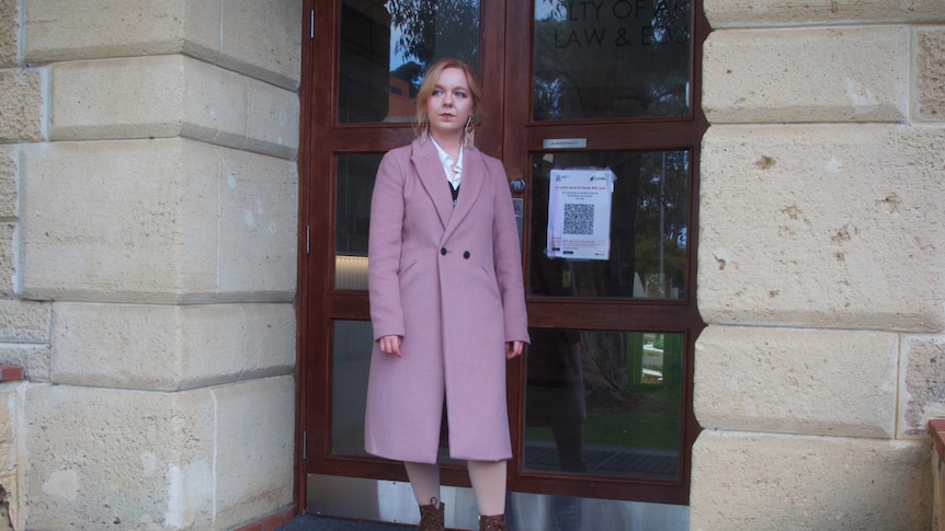 A young woman in a lilac jacket stands on the steps of a building