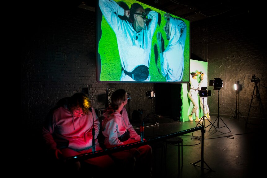 Two people look up at a large screen in a theatre space. Streamed onto the screen are two people acting against a green screen.