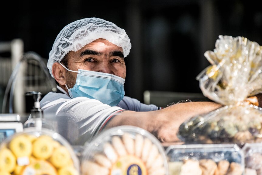 A shop attendant wearing a face mask and hair net leans on a deli counter displaying pastries and sweets in Liverpool.