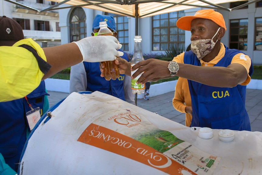 a man in a mask and blue vest hands another man a bottle containing a brown liquid