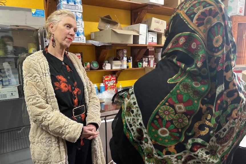 Gina standing with her hands clasped together, listening to a woman wearing a headscarf speaking in the community centre.