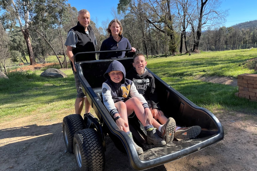 Woman and man stand with two sons in a wheelbarrow in a green clearing in the woods.