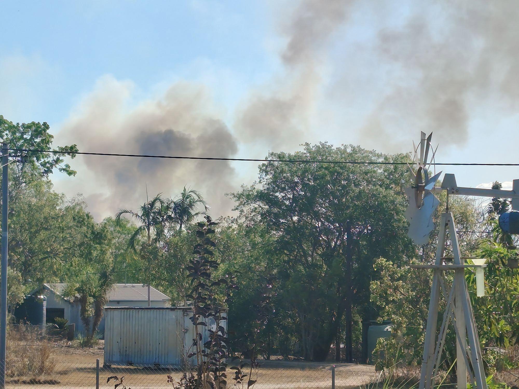 Plumes of bushfire smoke fill the air over a rural property, a windmill stands in the yard.