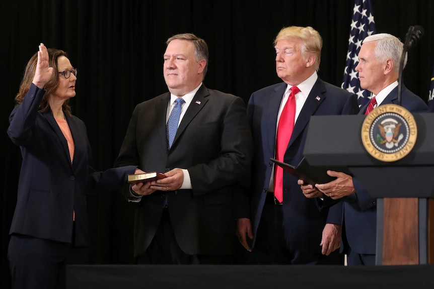 Gina Haspel raises one of her hand and has the other on the bible. Mike Pence, Donald Trump and Mike Pompeo are standing nearby.