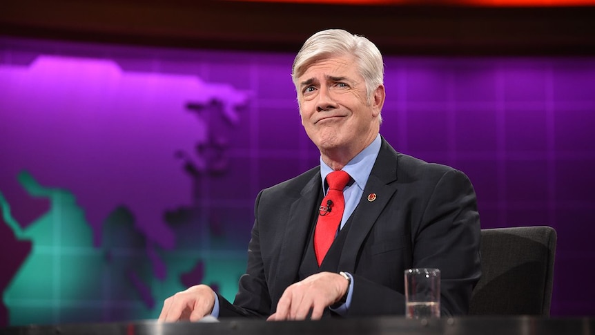 Shaun Micallef sitting at a desk, looking sideways and smiling.