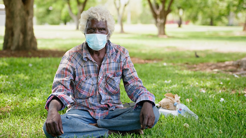 An Aboriginal man sitting cross-legged on a patch of grass, wearing a surgical face mask. 