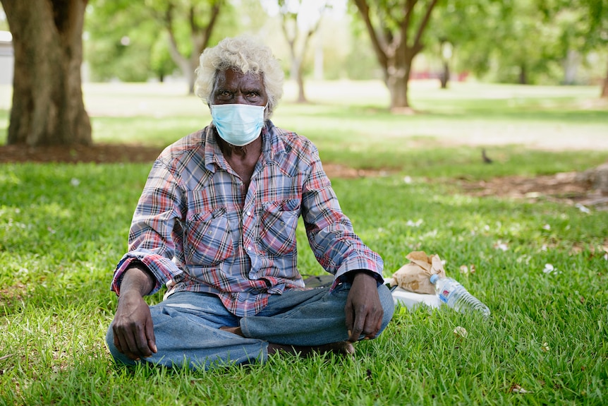 An Aboriginal man sitting cross-legged on a patch of grass, wearing a surgical face mask. 