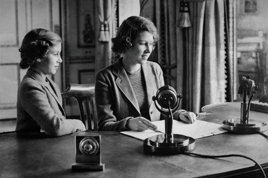 A young Queen Elizabeth speaking into a microphone on a desk while little Princess Margaret looks on