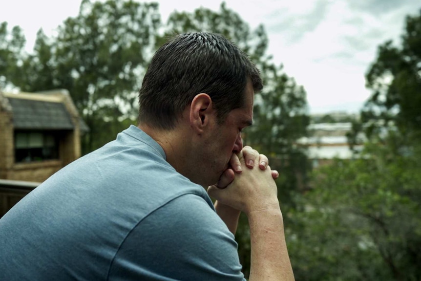 Man with black hair in blue t-shirt stands on balcony with head resting on hands with brick building and trees in background.