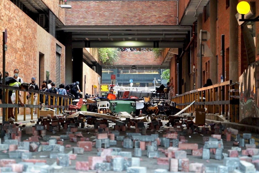 Bricks are laid on the ground while chairs and tables are piled into a barricade. Students walk nearby.