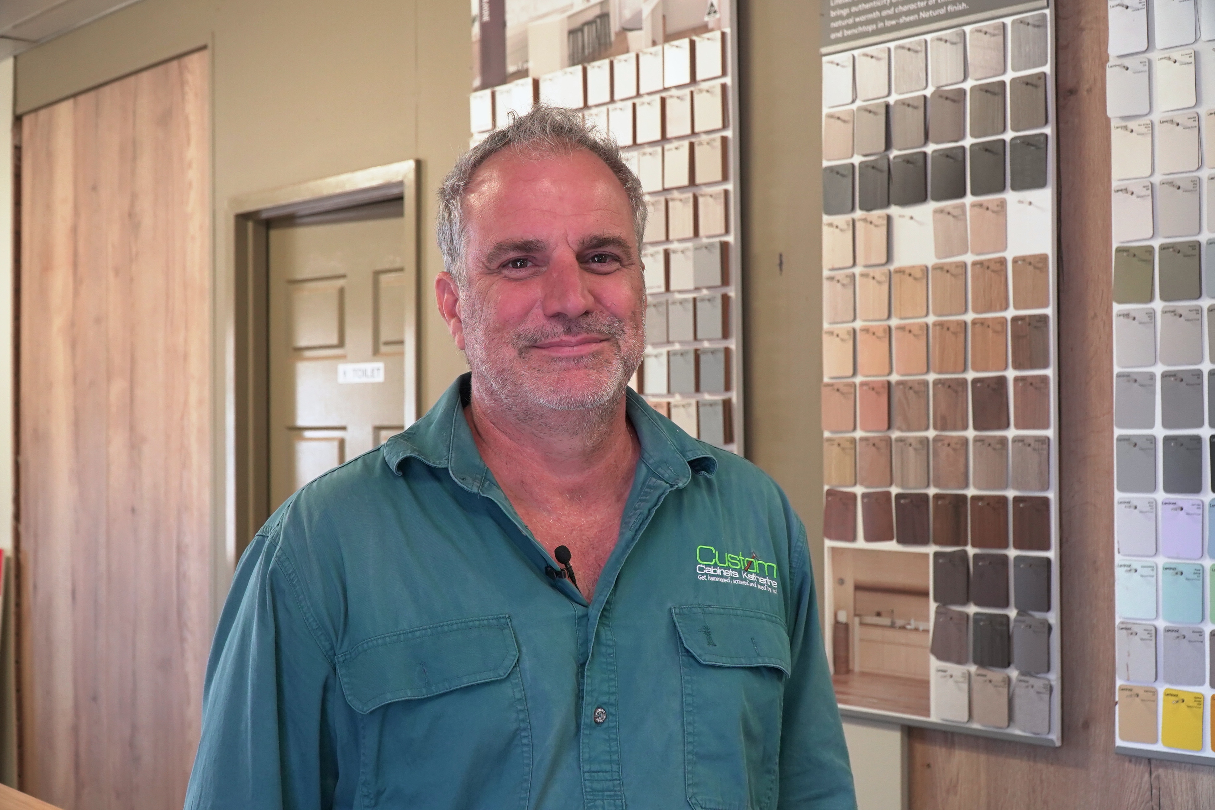 White man standing in a shop with sample wood behind him. He's wearing a green shirt.