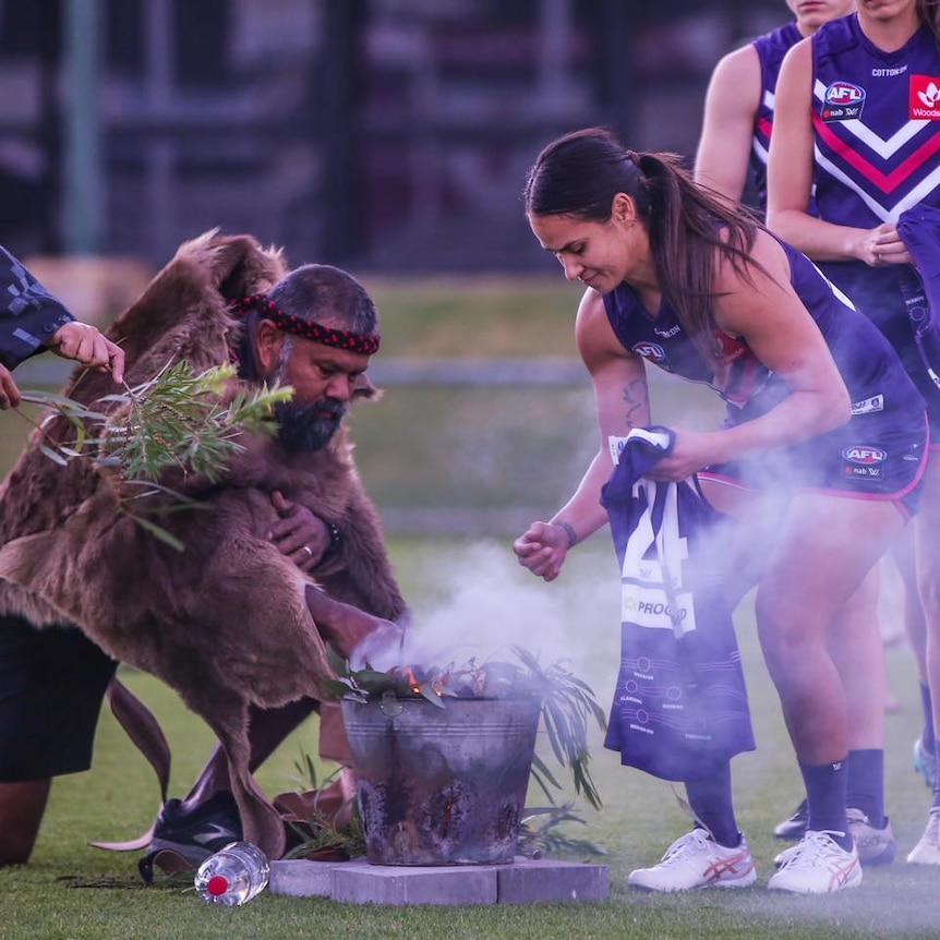 Indigenous man leaning over a traditional smoking ceremony with indigenous AFLW player wearing purple Fremantle Dockers uniform