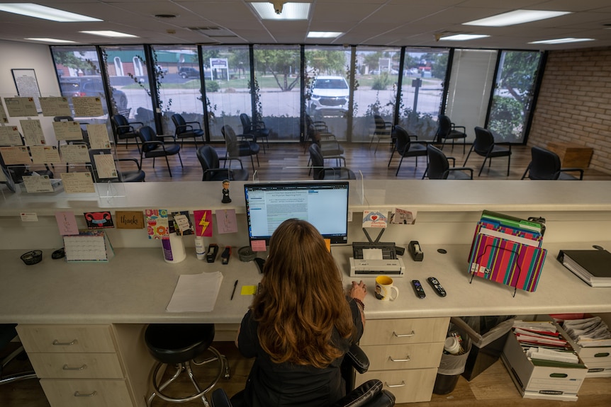 A woman sits behind a reception desk looking out onto an empty waiting room. The desk is covered with post cards