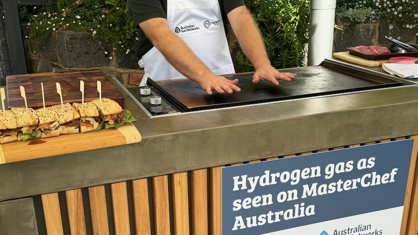 A person wearing a white apron holds their open palms slightly above a barbecue checking its heat.