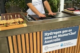 A person wearing a white apron holds their open palms slightly above a barbecue checking its heat.