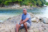 A Fijian man sits on a rock in front of water with trees on an island in the background.