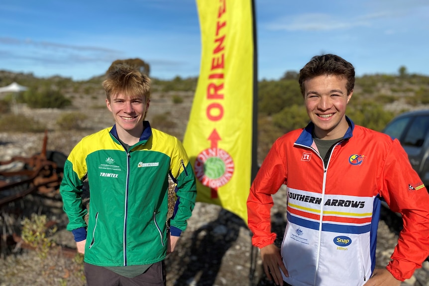 Guy on left in green and gold jacket, guy on right in red and white SA jacket, in front of yellow orienteering sign.