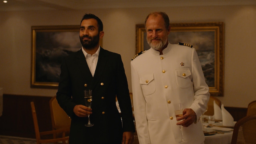 A young man in dark suit and an older man with beard in white captain's uniform holding glasses of sparkling and leaning 