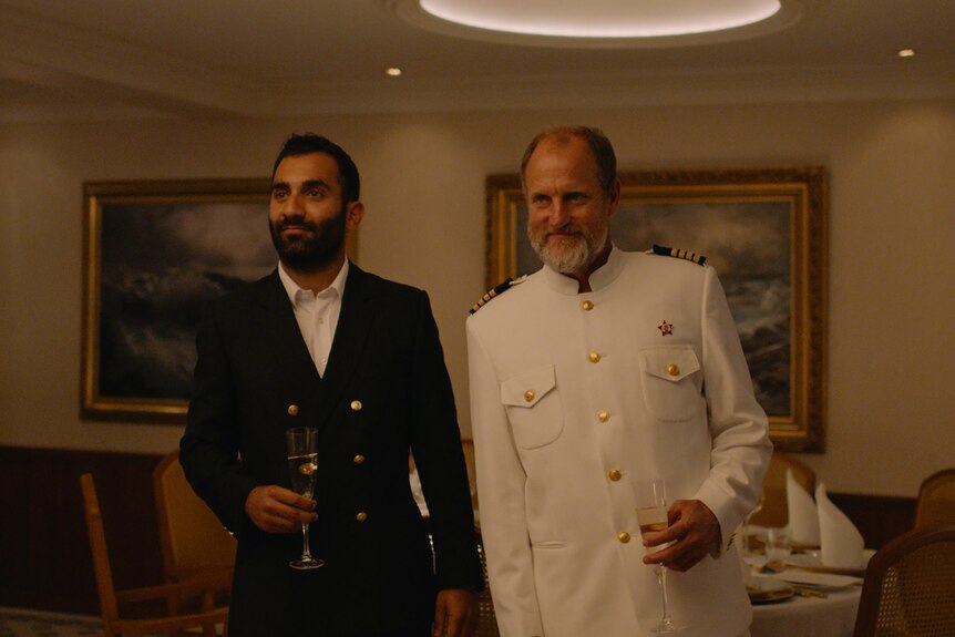 A young man in dark suit and an older man with beard in white captain's uniform holding glasses of sparkling and leaning 