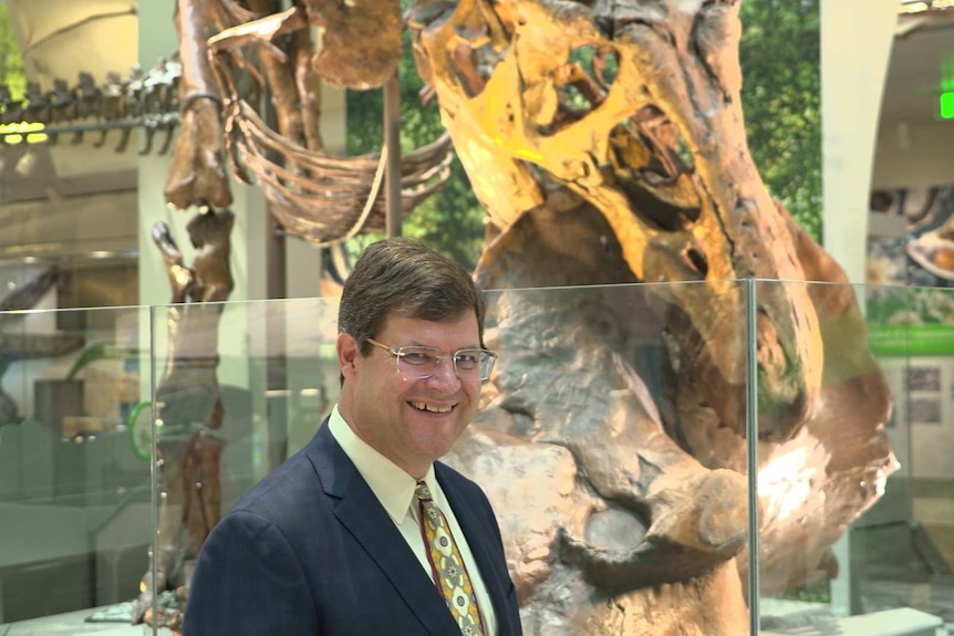 A man smiles while standing in front of a dinosaur fossil