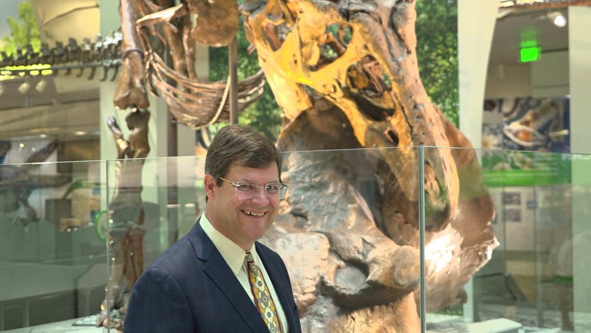 A man smiles while standing in front of a dinosaur fossil