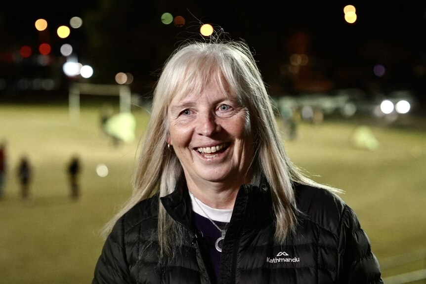 A middle-aged white woman with long blonde hair. She is in front of a suburban football pitch at night, lit by flood lights