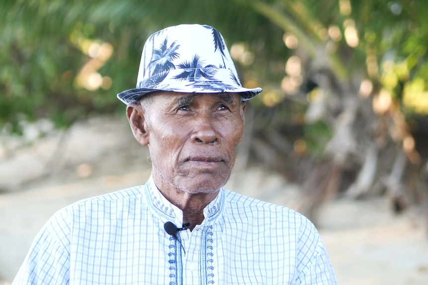 An older man wearing a hat with tropical patterns in front of a tropical green setting.