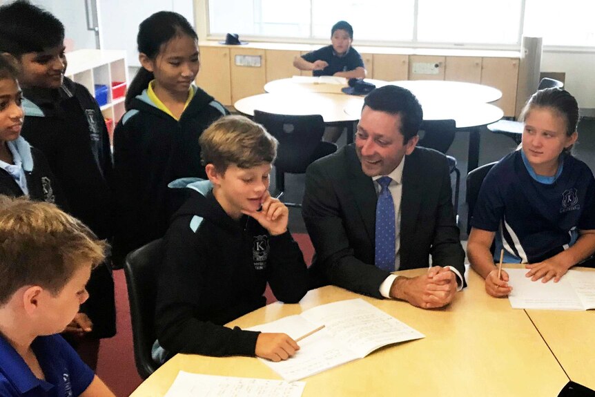 Matthew Guy at a table in a school classroom talking to kids.