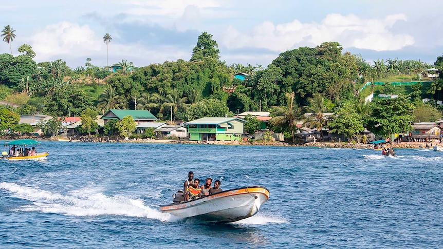 local people in a small motor boat on bright blue water in front of a colourful, modest town