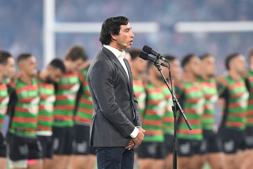 former nrl player jonathan thurston speaks into a microphone with a football team behind him. he is wearing a grey suit