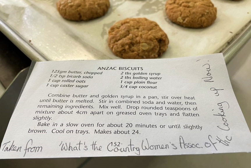 A printed-out recipe for Anzac biscuits with some handwritted notes on it, with biscuits in the background.
