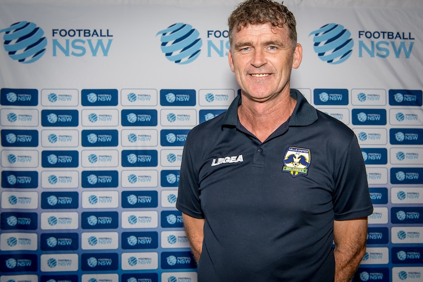 Alex Tobin, wearing a Hills United polo, stands in front of a Football NSW backdrop