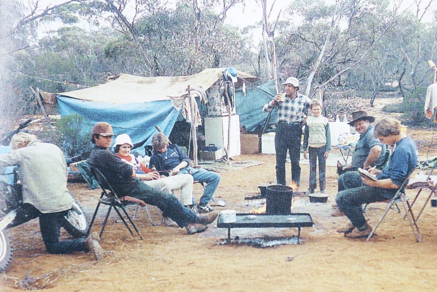 A campsite in the bush with adults and children on chairs around a camp oven, a makeshift tent in the background