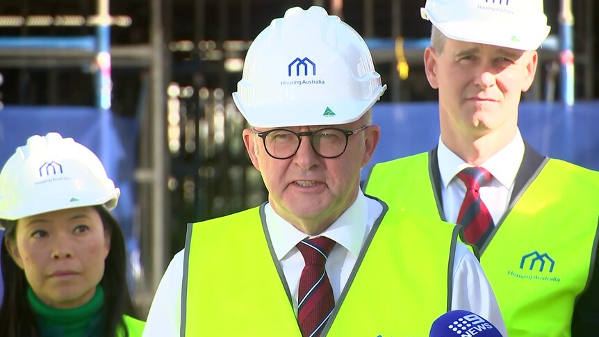 Albanese wearing a white hard hat and yellow vest flanked by two others in the same outfit
