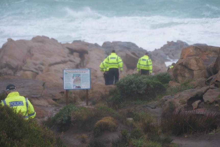 Three police searching rocks near the ocean with large swell in the background.