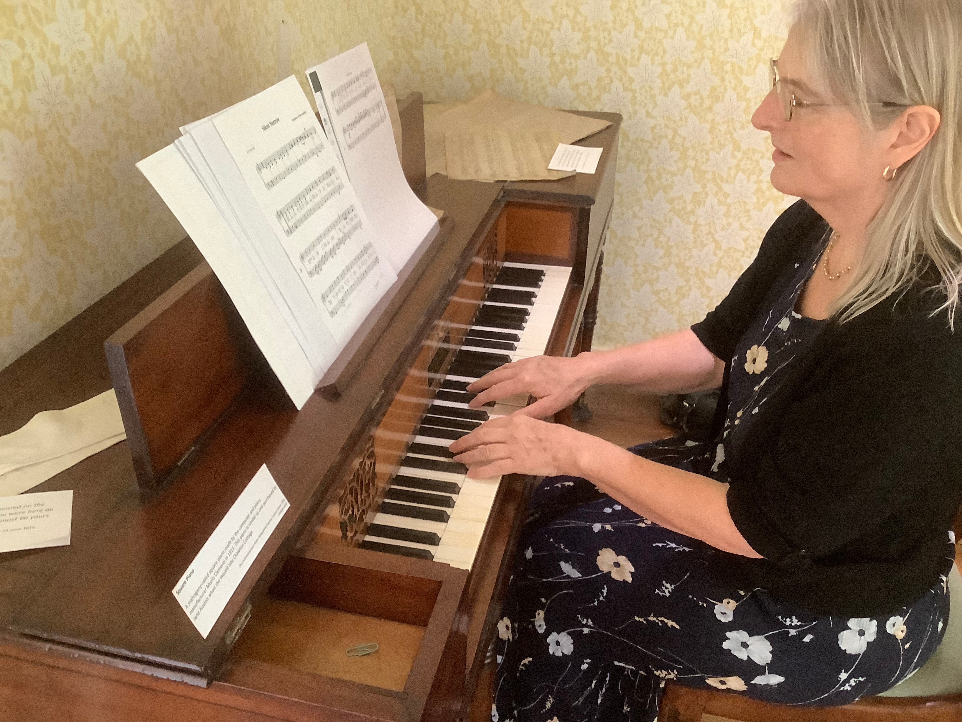 Gillian Dooley está sentada tocando um antigo piano quadrado de madeira. A partitura está na estante de partitura e espalhada pela parte superior do piano.