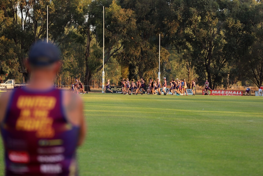Man in foreground looking on at large group of young men running around country football oval at dusk