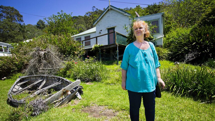 Marion Rae stands in front of a rundown cottage surrounded by bushland.