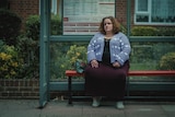 A woman in a blue cardigan and purple skirt sits at a bus stop