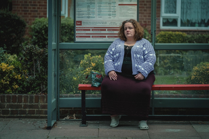 A woman in a blue cardigan and purple skirt sits at a bus stop