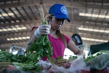 A woman holds a bunch of coriander above other produce at and undercover marketplace.
