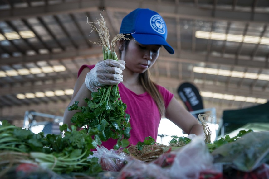 A woman holds a bunch of coriander above other produce at and undercover marketplace.