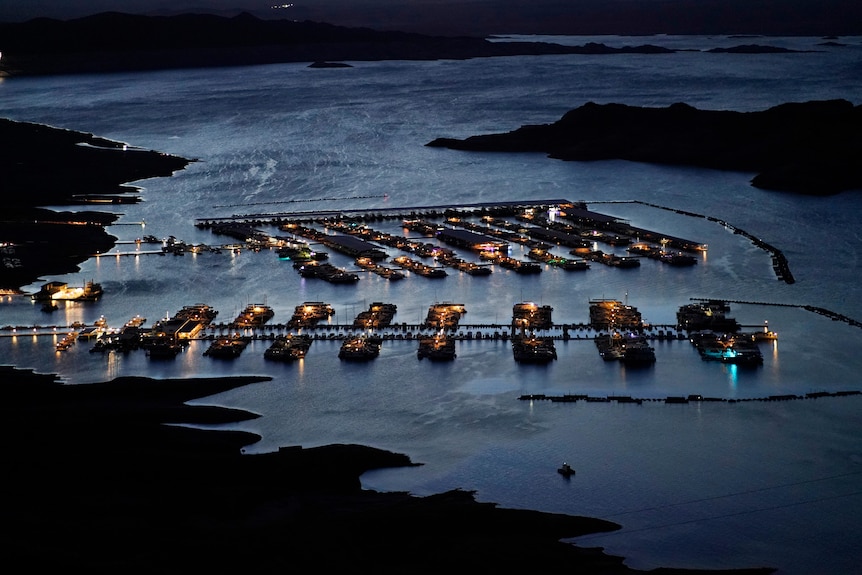 Elevated view of a boat harbour at night. Yellow lights are lit up on every boat moored at the dock. 