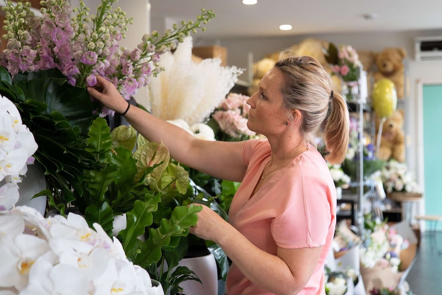 A blonde woman in profile reaching up to a wall of flowers