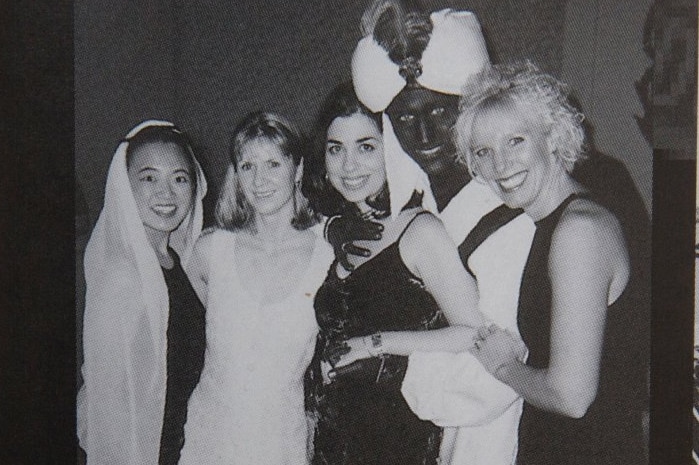 A black and white photo shows three women standing with Justin Trudeau, who is wearing dark makeup and an Arabian-style costume.