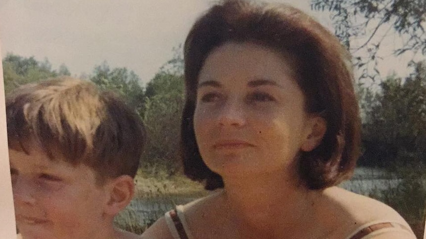 Old photo of young blonde boy sitting against a woman with short brown hair, who is subtly smiling. Both look off to side.