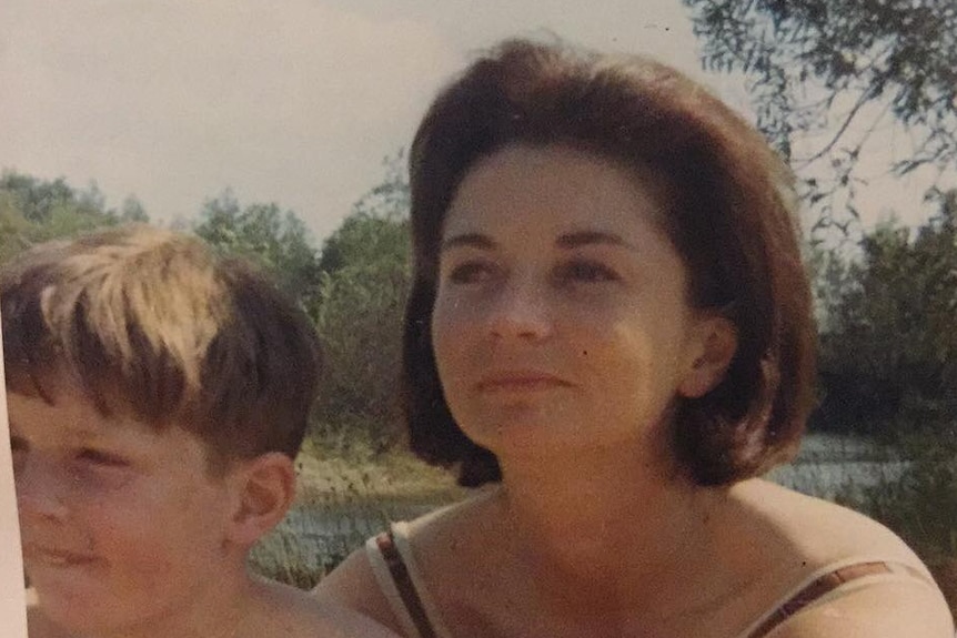 Old photo of young blonde boy sitting against a woman with short brown hair, who is subtly smiling. Both look off to side.