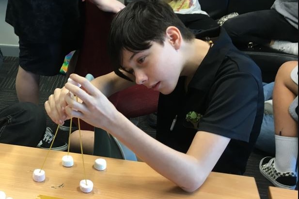 Riley, who has short hair and wearing black, hunched at a school desk holding 4 sticks together at the topwith marshmallow bases